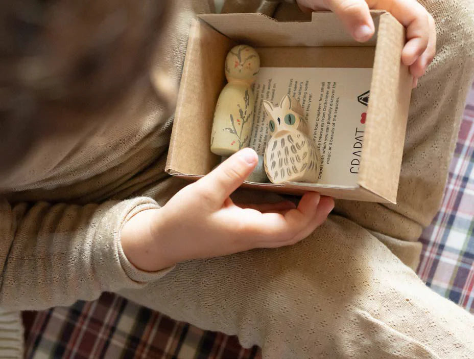 Person holding a small cardboard box with wooden figurines and a book inside.